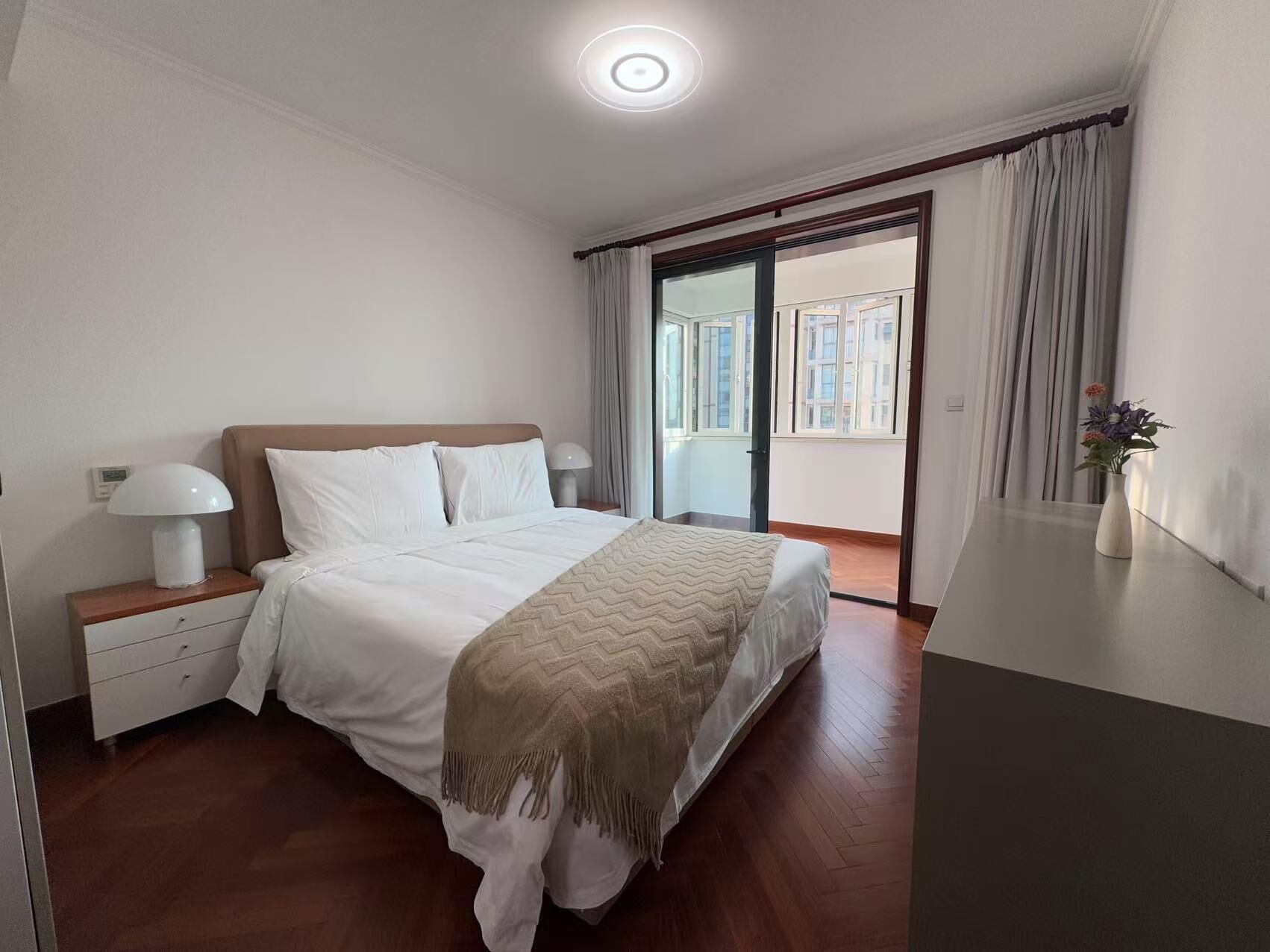 A bedroom with a bed, bedside tables, a lamp, a window with curtains, and a view of buildings.