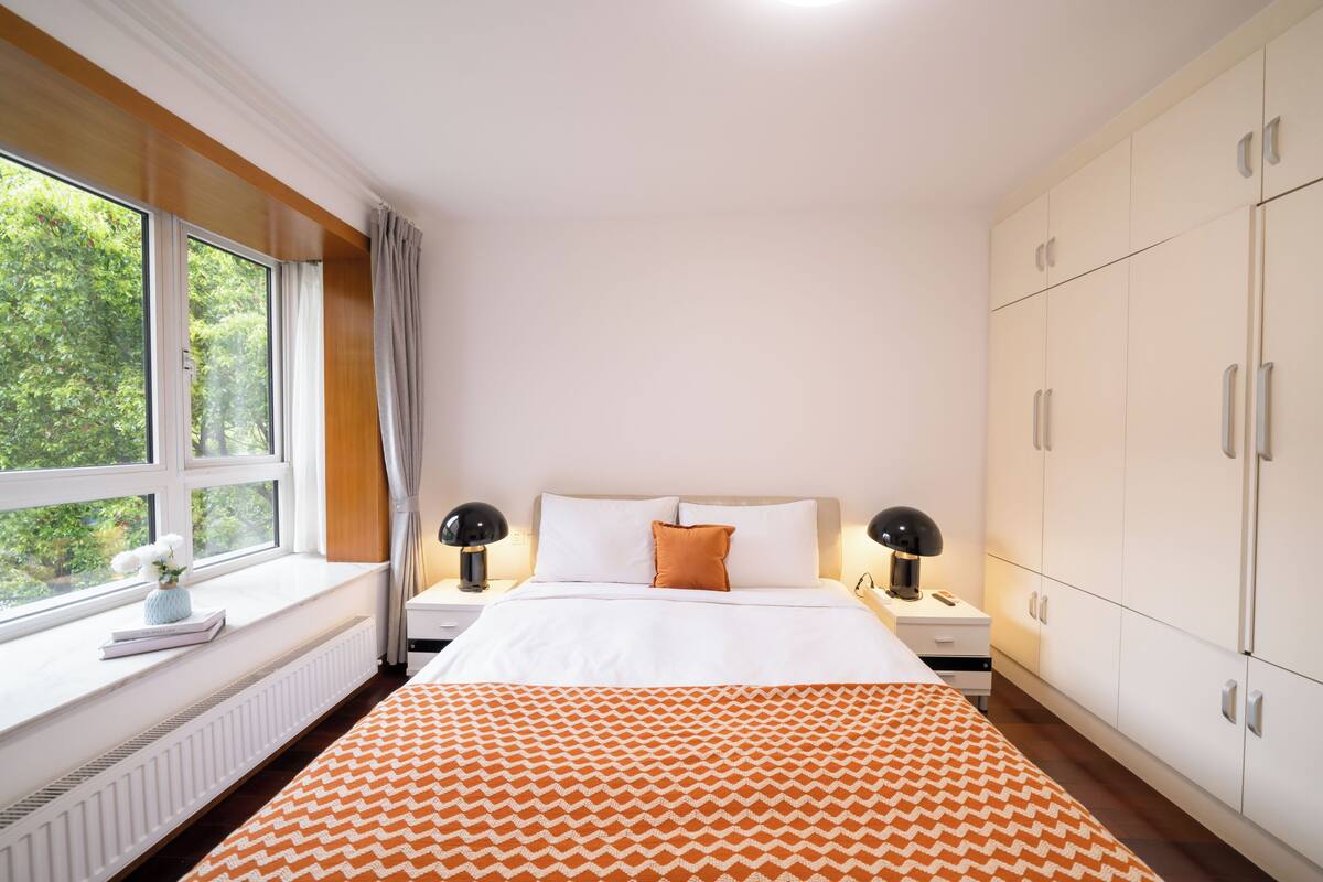 Un dormitorio con un ventanal grande, una cama con ropa de cama blanca, un edredón naranja estampado y un armario.