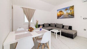 Ein modernes Wohnzimmer mit einem weißen Sofa, einem Esstisch mit Stühlen, einem Gemälde, das eine Strandansicht zeigt, und einem Fenster mit Vorhang.