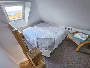 A small, single-room accommodation with a bed, a small table, a chair, a window with a view of the sea, and a power outlet on the wall.