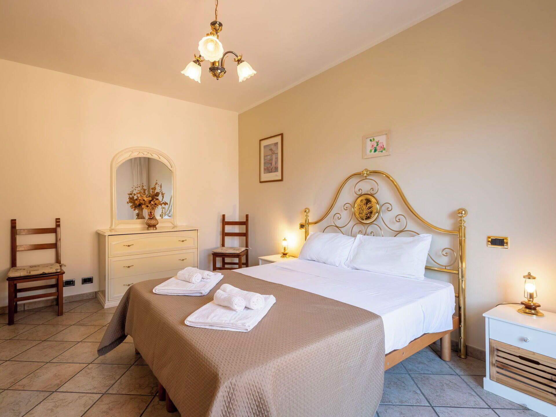 A bedroom with a bed, a dresser, a chair, a nightstand, and a chandelier.