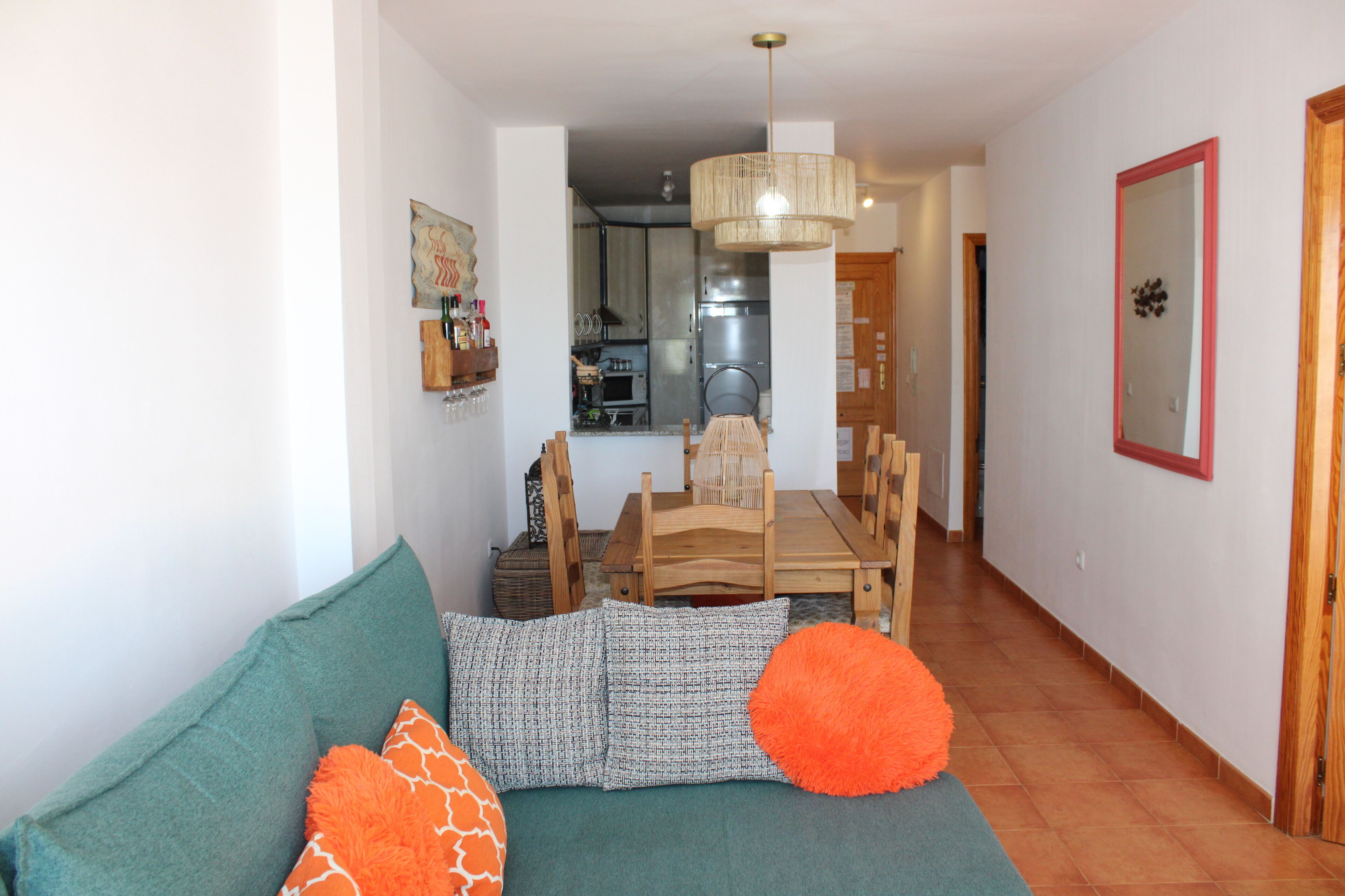 A living room with a sofa, pillows, a dining table, chairs, a hanging light fixture, and a kitchen in the background.