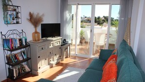 Ein Wohnzimmer mit einer Couch, einem Bücherregal, einem Flachbildfernseher und einer Glastür, die auf eine Terrasse im Freien führt.