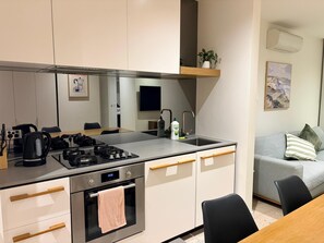 A modern kitchen with a gas stove, oven, and a built-in microwave. There is a sink with a faucet, a coffee maker, and a bottle of dish soap. A gray sofa and a plant are visible in the background. - New 2BR Inner-City Apartment | Free Parking | Near Marvel Stadium (West Melbourne)