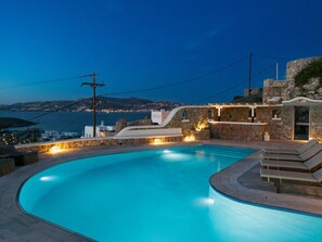 A swimming pool with a curved edge, stone walls, and a view of the sea and distant buildings. - Patron 1 Pool Sunset Villa- Seven Bedrooms (Mikonos)
