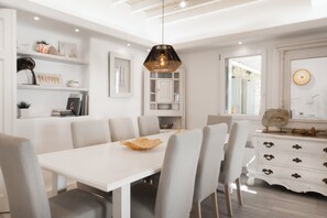 A dining area with a white table, grey upholstered chairs, a decorative hanging light, and a white cabinet with drawers. - Patron 1 Pool Sunset Villa- Seven Bedrooms (Mikonos)