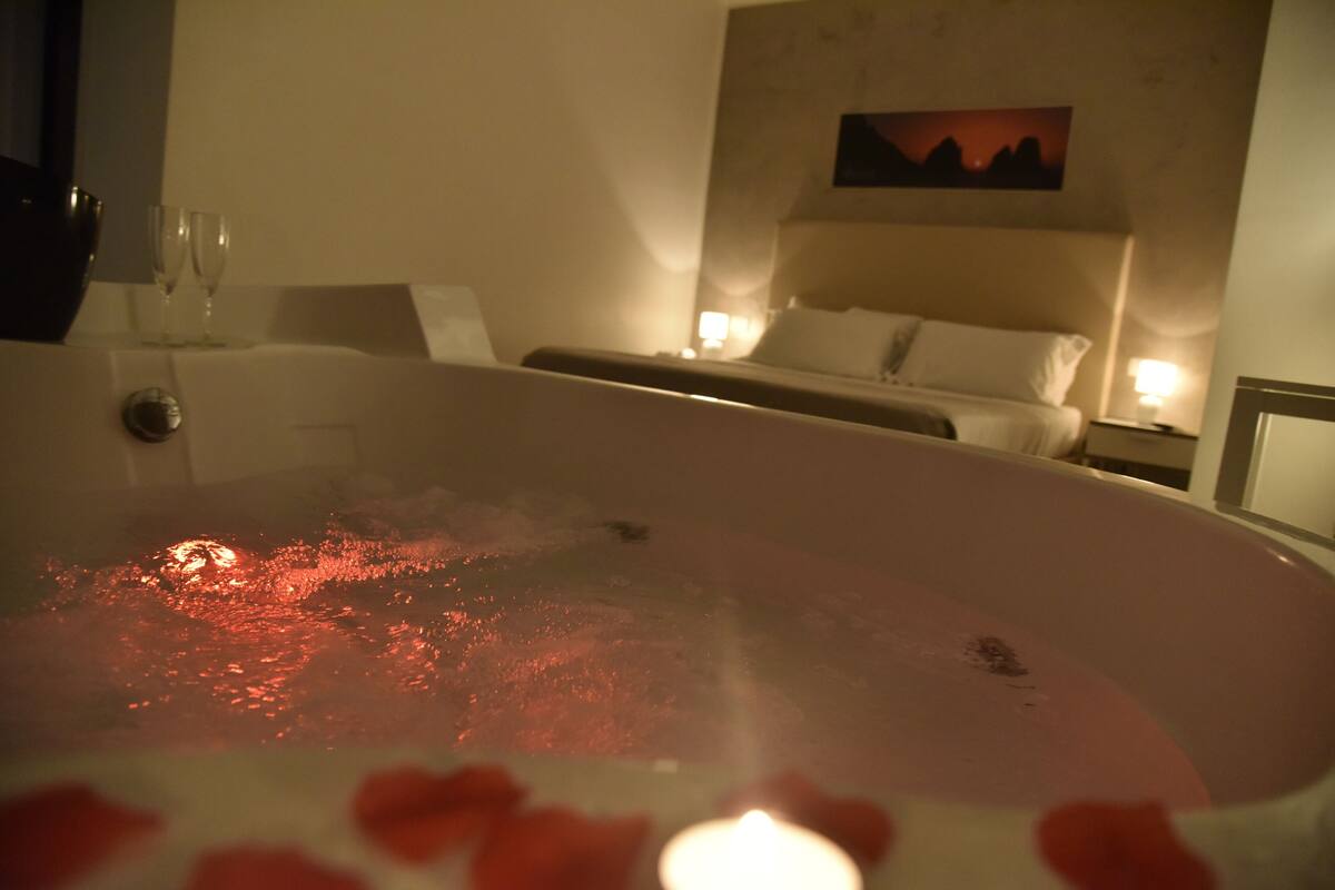 A room with a hot tub, a bed, a painting on the wall, and two bedside lamps.