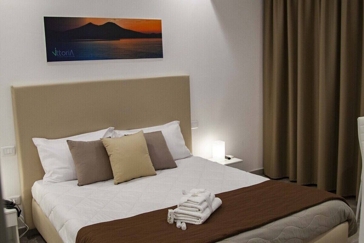 A hotel room with a bed, a nightstand, a wall-mounted air conditioner, a painting, and a window with curtains.