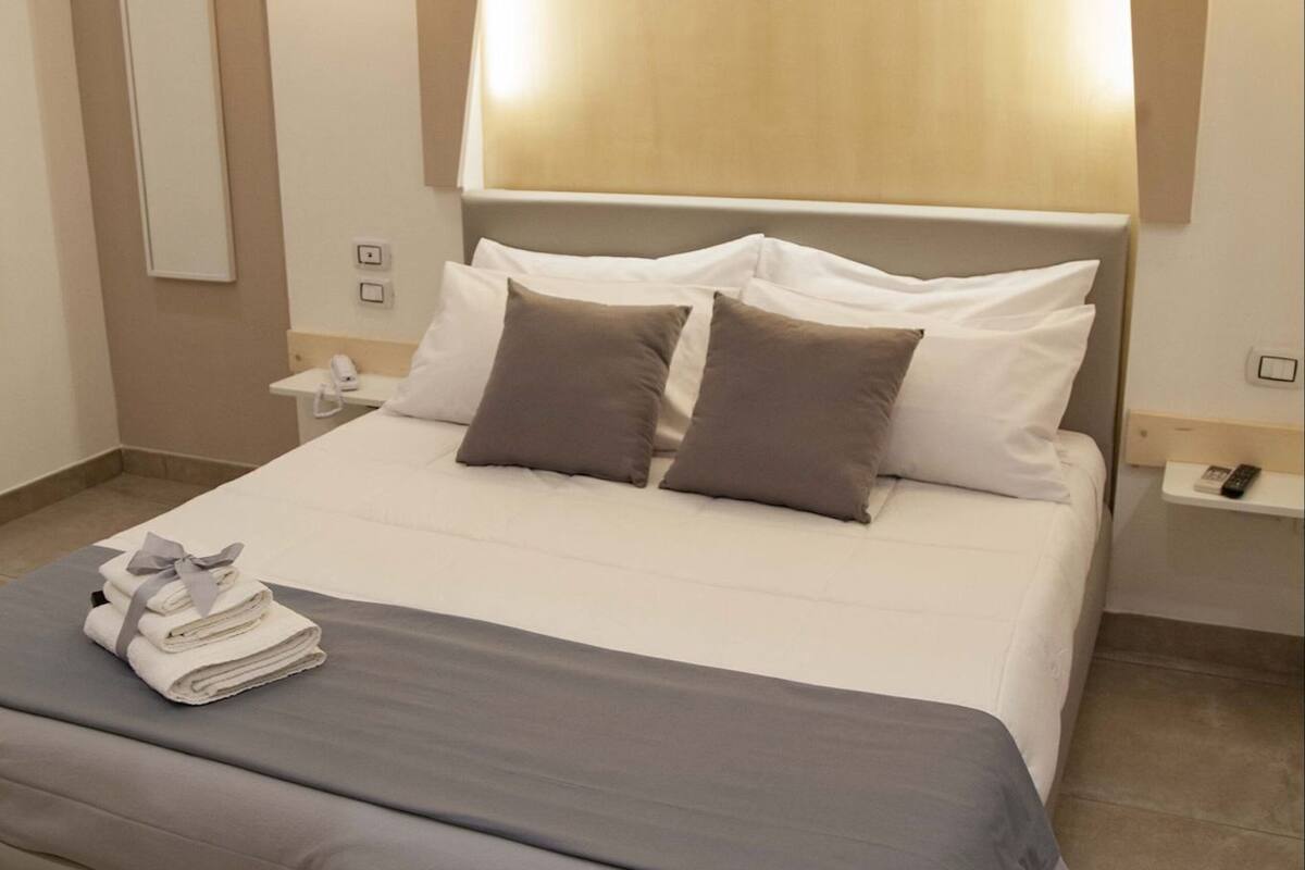A neatly made bed with a grey bedspread, a headboard with a light fixture, a bedside table with a phone, and a wall-mounted light.