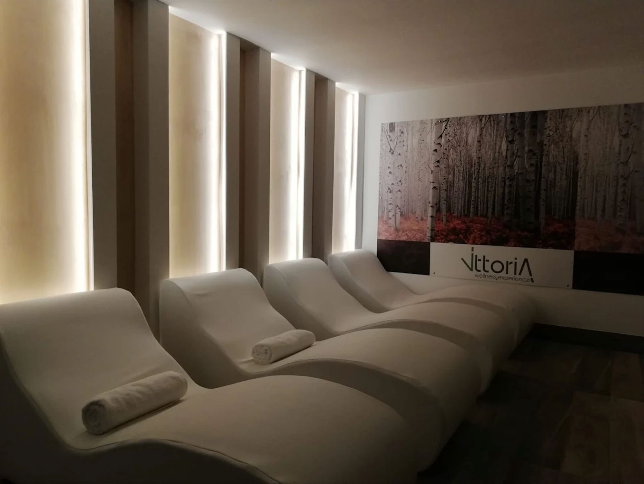 A row of white lounge chairs with towels, a large wall mural, and a sign reading 'Vittoria wellness experience'.