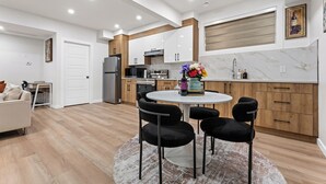 A modern kitchen with white cabinets, a dining area with a round table and chairs, and a living room with a sofa. - 2BR Near YEG Airport | King Bed + Mini Golf! (Nisku)