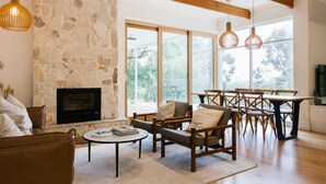 A spacious living room with a stone fireplace, wooden furniture, large windows, and hanging pendant lights.
