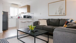 Een moderne woonkamer met een grijze bank, een bijzettafeltje met een schaal fruit, en een keuken op de achtergrond.