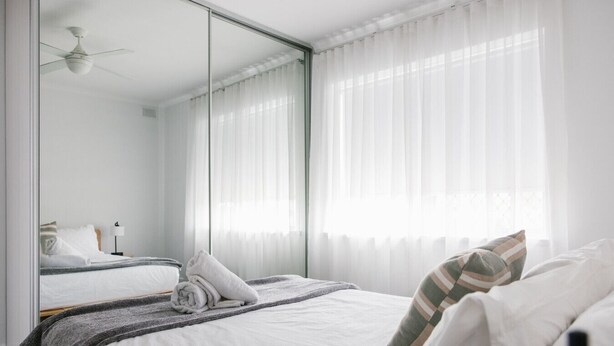 Een moderne slaapkamer met een grote spiegel, een bed met witte lakens, een plafondventilator en dunne gordijnen.