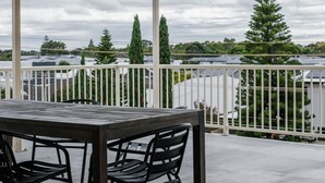 Een overdekt terras met een tafel, stoelen, een balustrade en uitzicht op bomen en gebouwen.