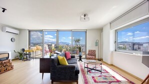 A modern living room with a sofa, a coffee table, and a view of a cityscape through large windows. - Amazing Acropolis Views Penthouse in Buzzy Gazi (Athens)