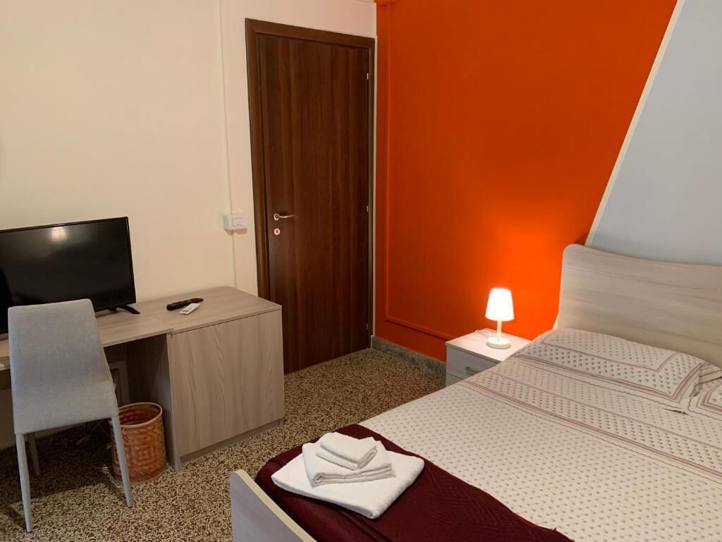 A hotel room with a bed, a desk with a computer, a chair, a television, and a nightstand with a lamp.