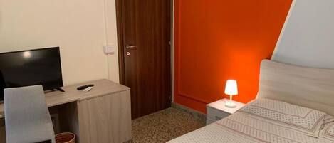 A hotel room with a bed, a desk with a computer, a chair, a television, and a nightstand with a lamp.