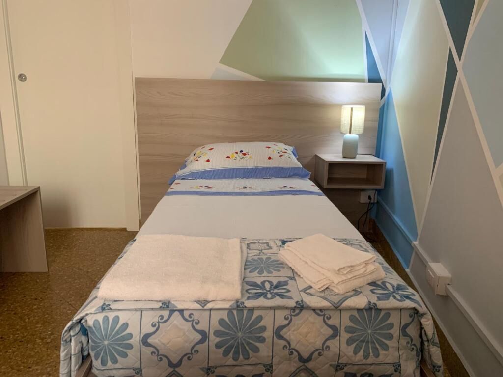 A bedroom with a bed, a bedside table with a lamp, a wall-mounted shelf, and a door.