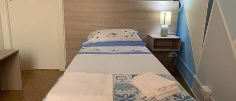 A bedroom with a bed, a bedside table with a lamp, a wall-mounted shelf, and a door.