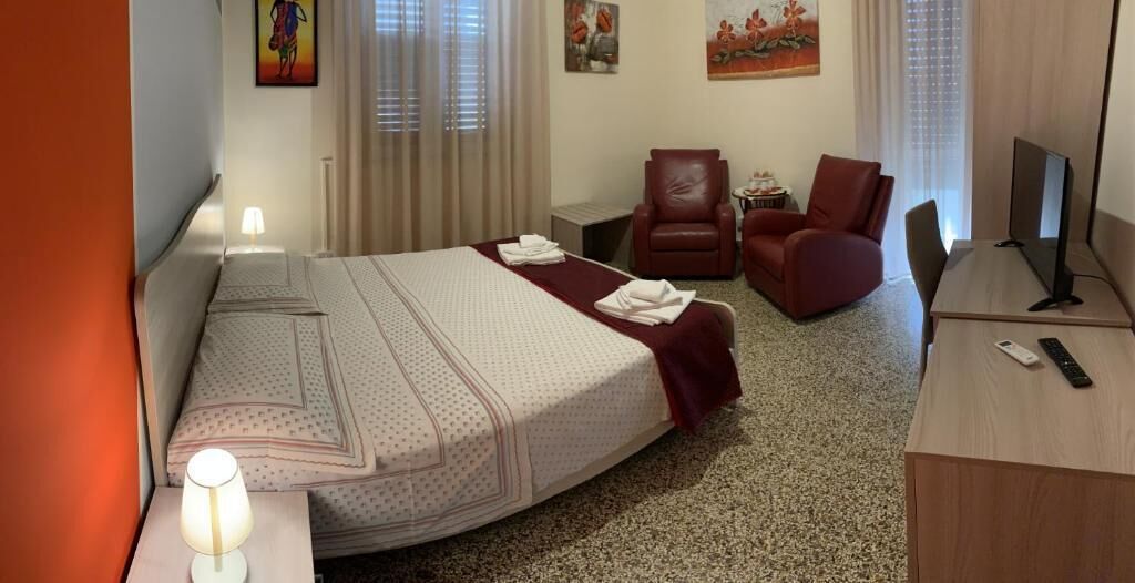 Ein Hotelzimmer mit einem Bett, zwei Stühlen, einem Schreibtisch mit einem Computer, einer Lampe und Bildern an den Wänden.