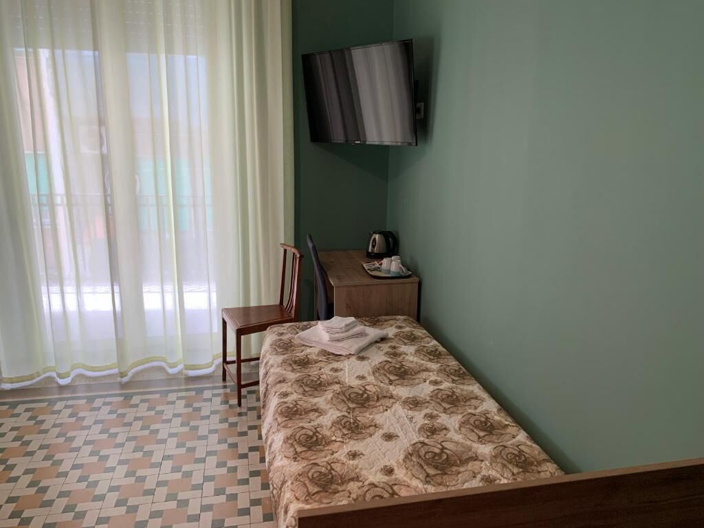 Ein Zimmer mit einem Bett, einem Schreibtisch, einem Stuhl, einem Fernseher und einem Fenster mit transparenten Vorhängen.