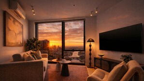 Ein modernes Wohnzimmer mit Sofa, Sesseln und Fernseher. Ein großes Fenster bietet Blick auf die Stadt bei Sonnenuntergang.
