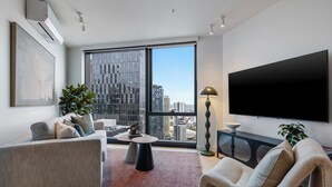 Ein modernes Wohnzimmer mit großem Fernseher, einer Couch, einem Sessel und einem großen Fenster mit Blick auf die Stadt.