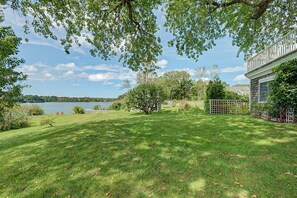 A grassy backyard with a view of a lake, a wooden fence, and a building with a white exterior. - Timeless charm at this unique waterfront compound, sleeping 14. (West Dennis)