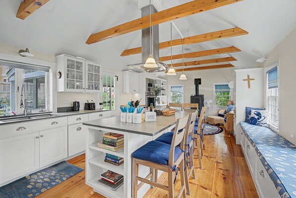 A modern kitchen with white cabinetry, a central island with bar stools, and a dining area with a blue upholstered bench. - Timeless charm at this unique waterfront compound, sleeping 8. (West Dennis)