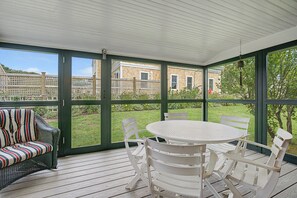 A covered patio with a round table and chairs, a wicker sofa, and a view of a wooden house and garden. - Timeless charm at this unique waterfront compound, sleeping 8. (West Dennis)