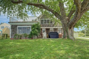 A two-story house with a large tree in the front yard, a covered grill, and a white fence. - Timeless charm at this unique waterfront compound, sleeping 8. (West Dennis)