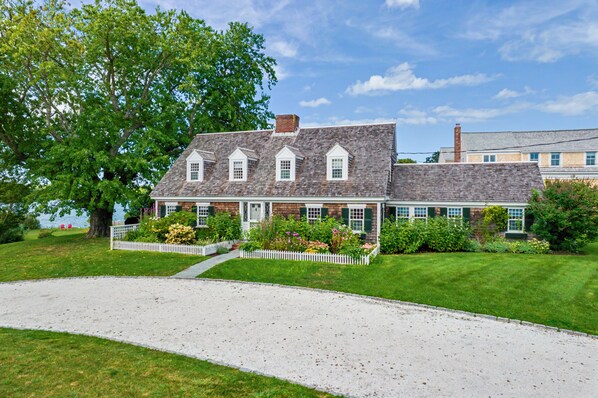 A two-story house with a white picket fence, a gravel driveway, and a well-maintained lawn. - Timeless charm at this unique waterfront compound, sleeping 8. (West Dennis)