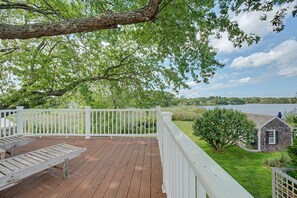 A wooden deck with white railings overlooking a lake, a grassy area, and a small building with green shutters. - Timeless charm at this unique waterfront compound, sleeping 8. (West Dennis)