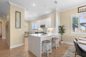 A modern kitchen with white cabinetry, a central island with stools, and a dining area with a table and chairs. - Corner Condo #141 with Ocean & Golf Course Views at Cinnamon Beach (Palm Coast)
