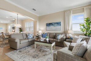 A spacious living room with a large window, a dining area, and a kitchen in the background. - Corner Condo #141 with Ocean & Golf Course Views at Cinnamon Beach (Palm Coast)