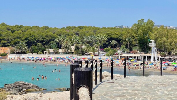 Ein Strand mit einem Kiesweg, der zum Wasser führt, umgeben von Sonnenschirmen und badenden Personen.