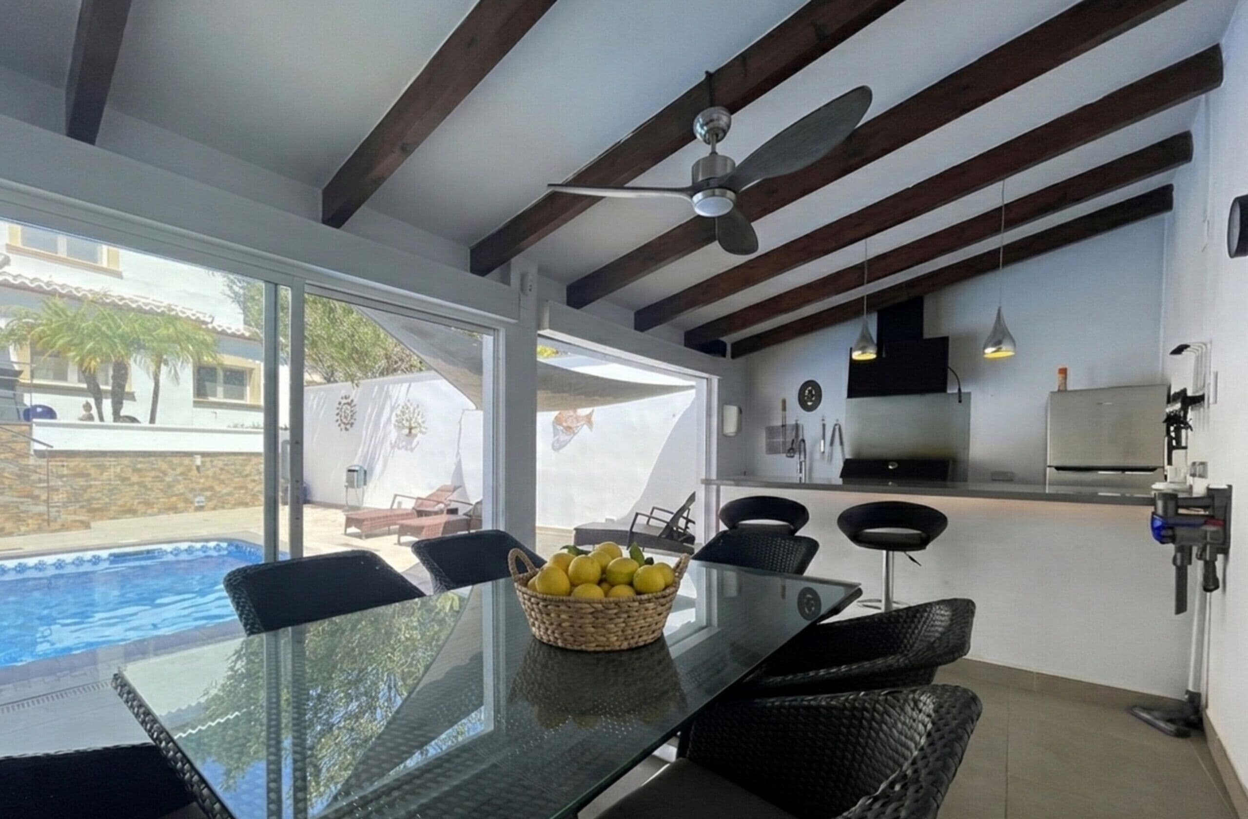 Una cocina moderna con una isla grande, un comedor con mesa de vidrio, y una piscina visible a través de puertas de cristal.