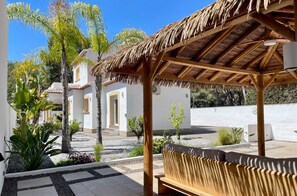 A covered outdoor seating area with a thatched roof, surrounded by plants and a building with a white facade. - Villa with private pool for 8 people – CASA SUSAN (Moraira)