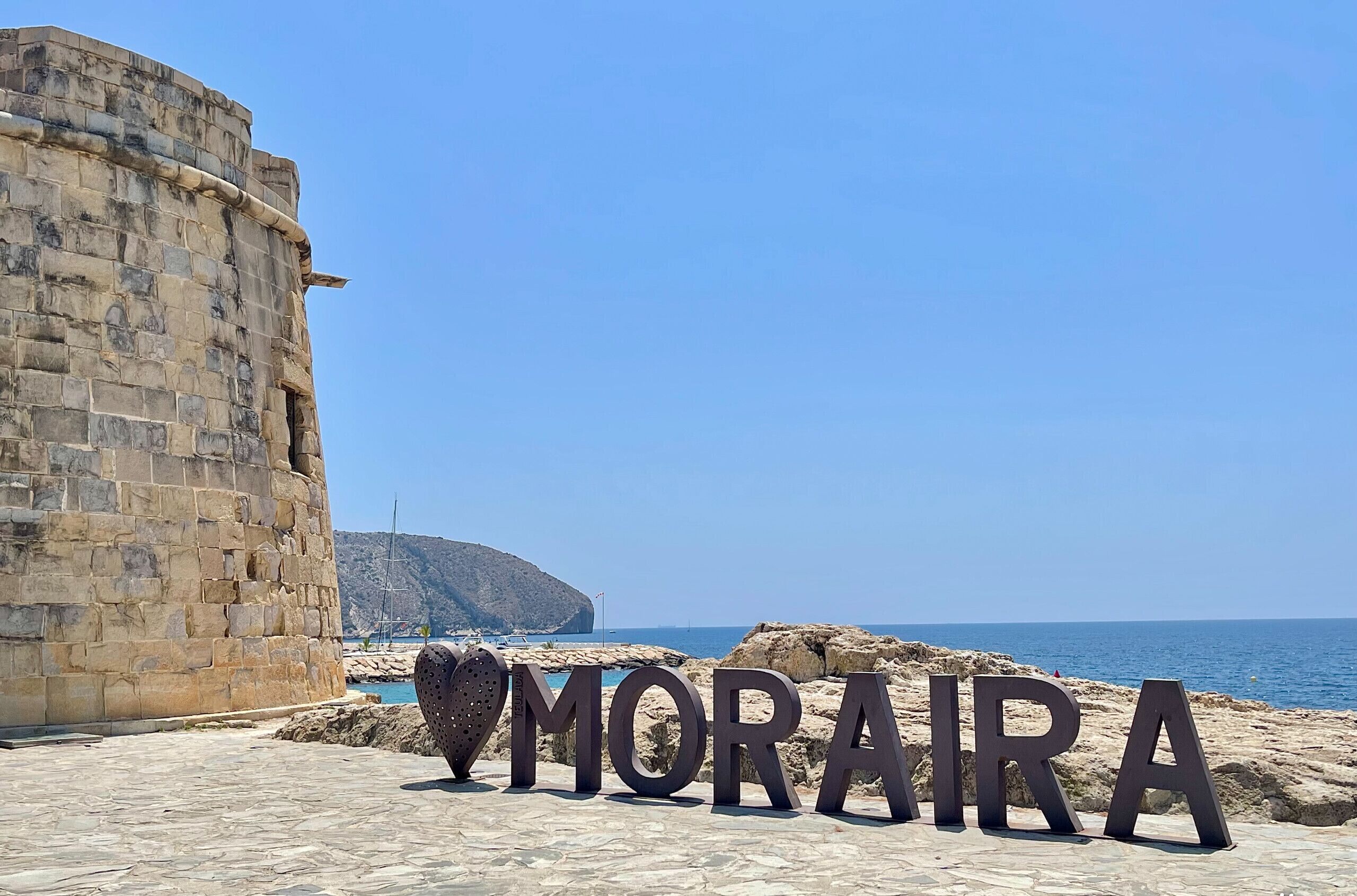 A stone wall and the word "MORIRA" in large letters on a paved area with a view of the sea and a hill in the background.