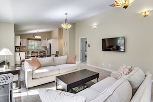 A living room with a sofa, coffee table, and a TV. There is a kitchen in the background with cabinets and a refrigerator.