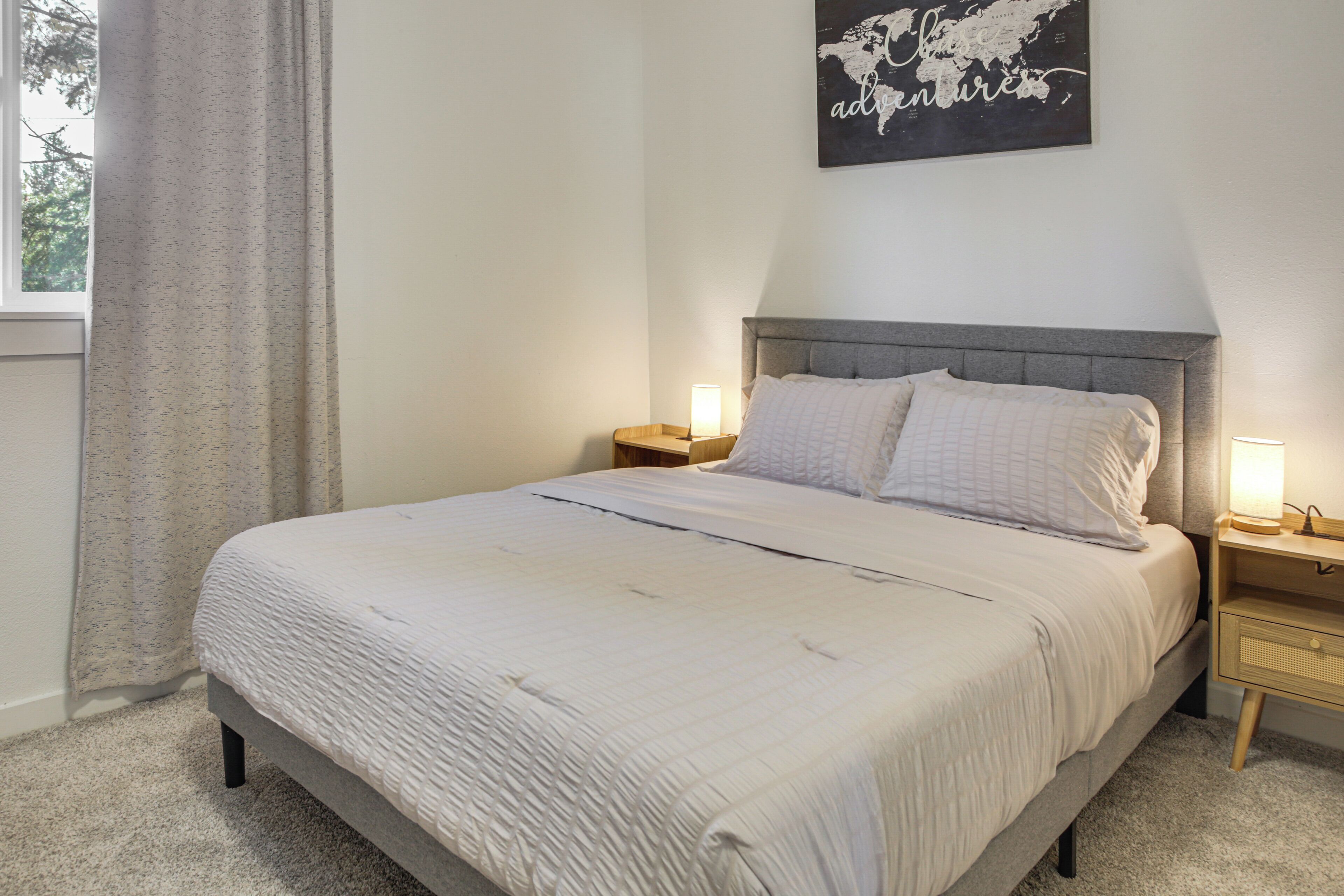 Een net opgemaakt bed met een grijs hoofdeinde, twee nachtkastjes met lampen, een wereldkaart aan de muur en een raam met gordijnen.