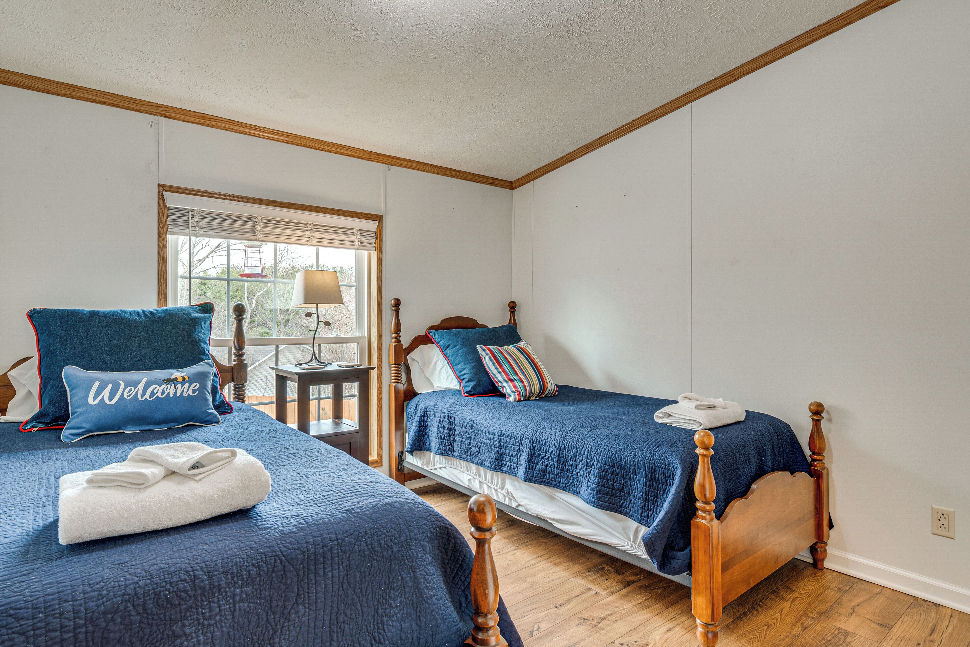 Et soveværelse med to senge, et vindue med skodder, et lille bord med en lampe og en pude med teksten 'Welcome'.