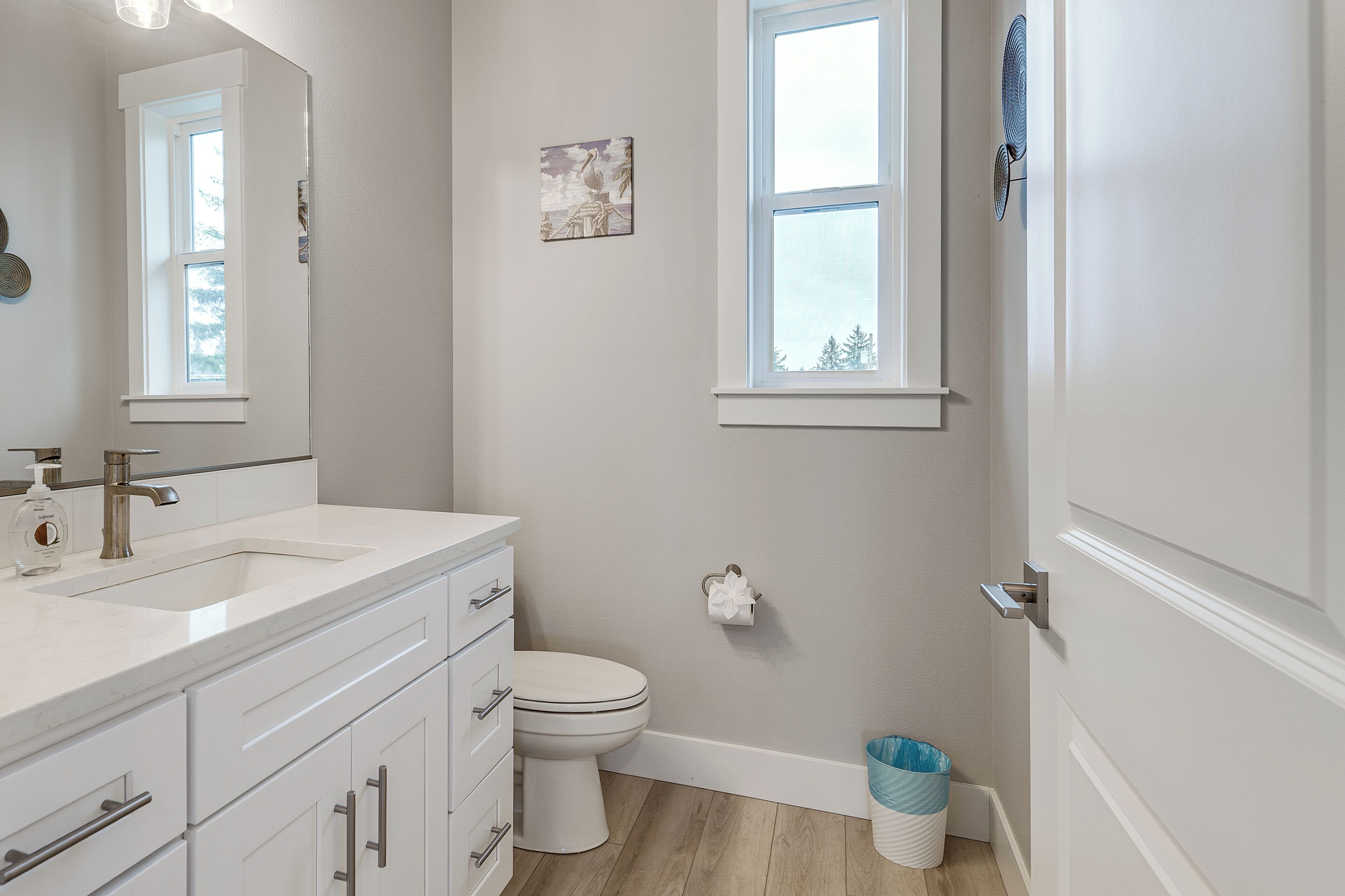A bathroom with a white sink, mirror, and toilet, a window, and a trash can.