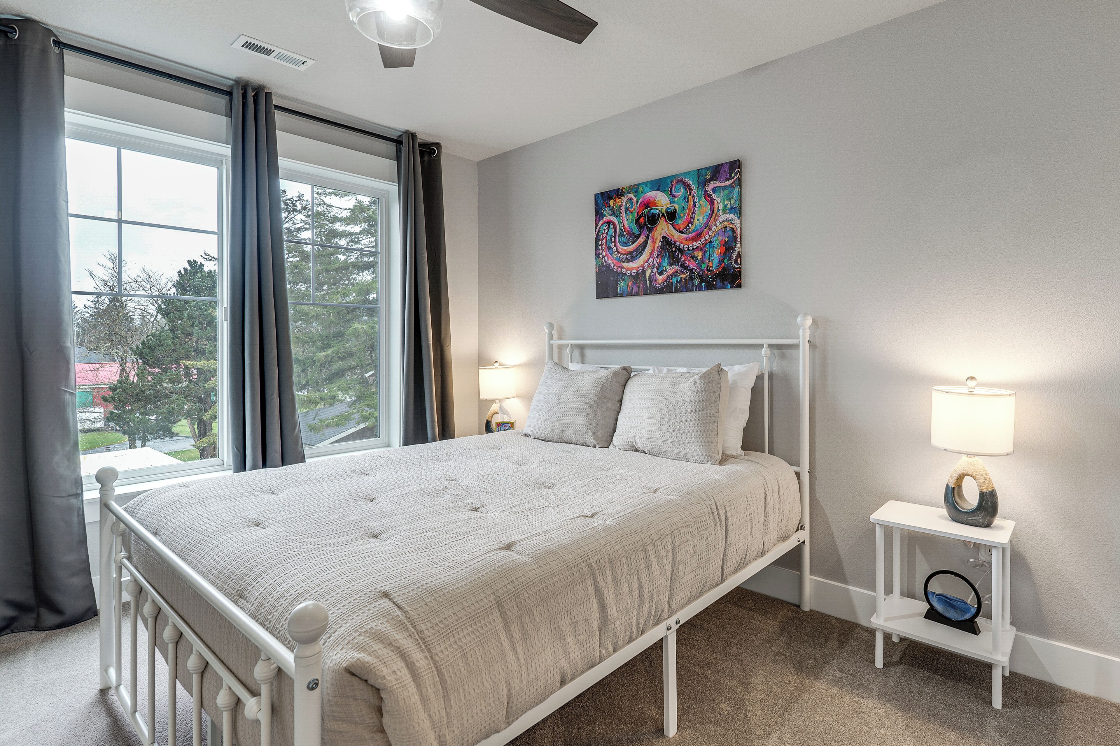 A bedroom with a bed, a window, a painting, and a bedside table with a lamp.