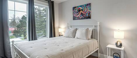 A bedroom with a bed, a window, a painting, and a bedside table with a lamp.
