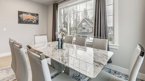 A dining room with a marble table, grey upholstered chairs, a painting of a fish, and a view of a house through large windows. - Walk to Marina! River-view Retreat in Hammond (Hammond)