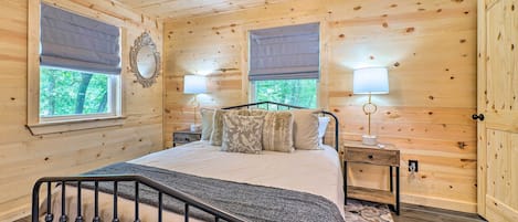 A wooden cabin bedroom with a bed, two bedside tables, and a window with a view of trees.