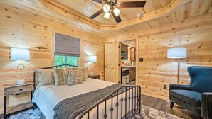 Une chambre dans une cabane en bois, avec un lit, des lampes de chevet, un ventilateur de plafond et un fauteuil bleu.