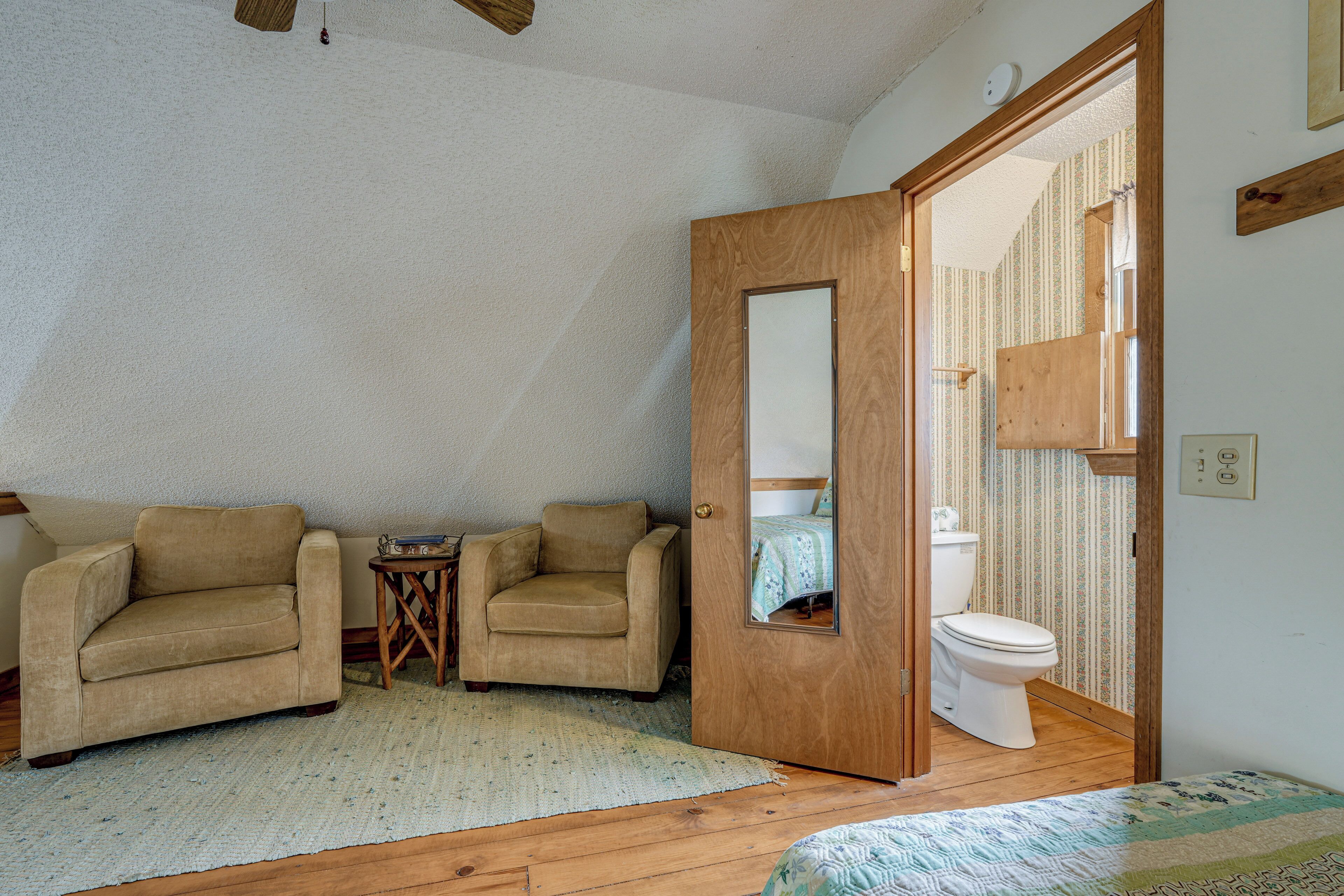 A bedroom with a wooden floor, two beige armchairs, a small wooden side table, a white door, and a visible bathroom with a toilet and shower.